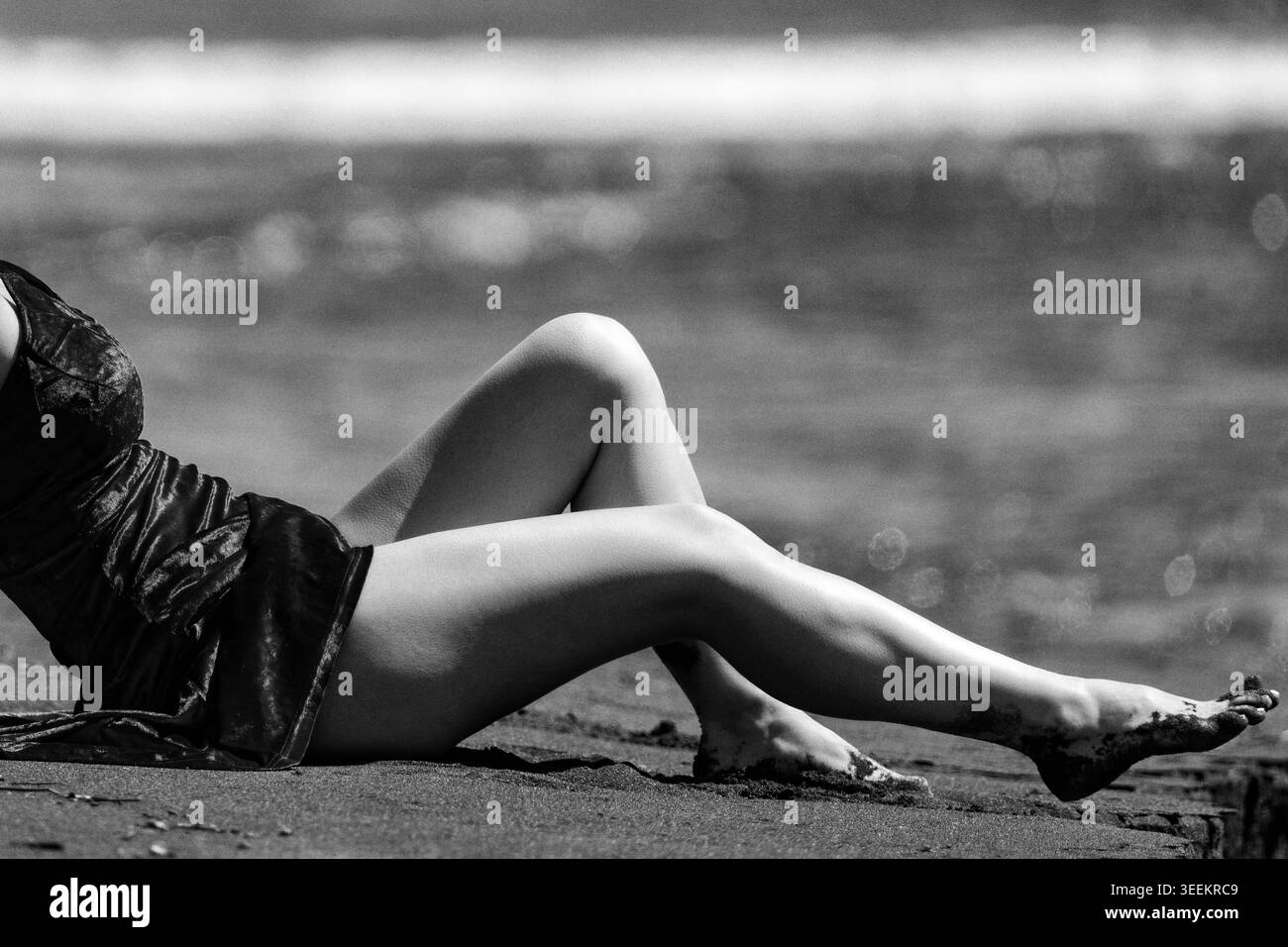Donna in abito che riposa sulla spiaggia durante il giorno di sole. Fotografia monocromatica di gambe femminili sulla sabbia. Scena marina in stile vintage con pellicola granulosa, Foto Stock
