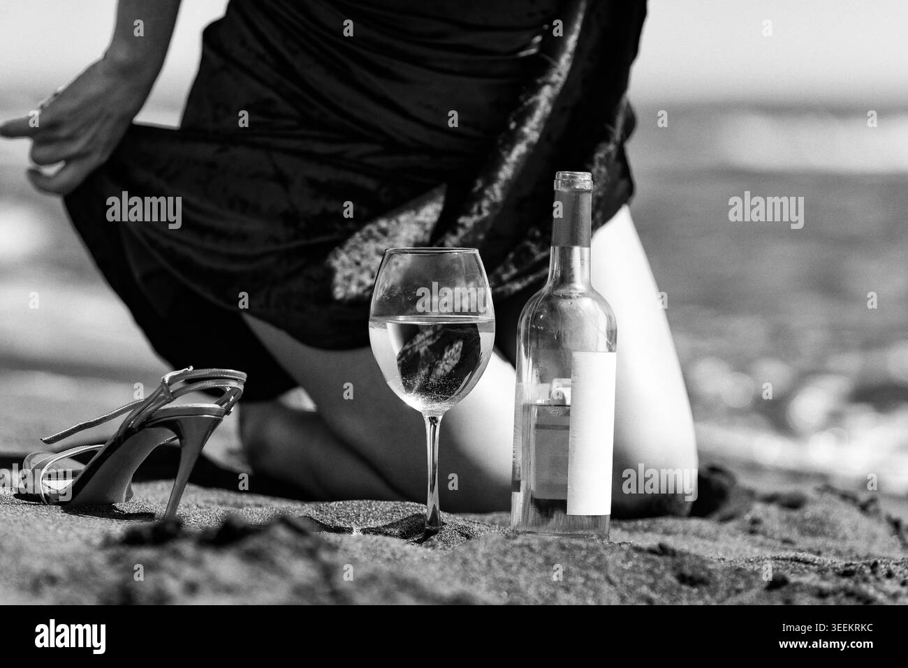 Donna inginocchiata sulla sabbia con bottiglia di vino e bicchiere sulla spiaggia. Serata romantica con scarpa vicino a bevande. In bianco e nero, stile cinematografico vintage con un Foto Stock