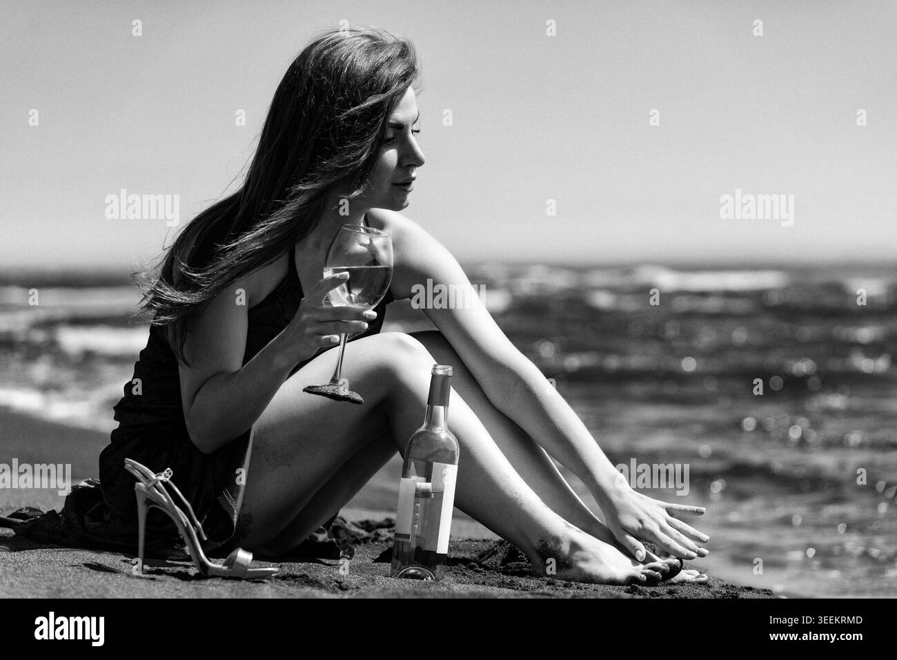 Donna seduta sulla spiaggia con un bicchiere di vino. Signora che ama le vacanze estive vicino all'oceano. Rilassatevi con bevande alcoliche. Concetto romantico in nero e. Foto Stock