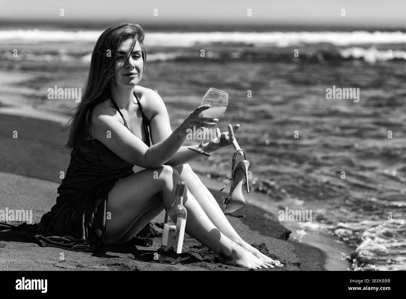 Donna seduta sulla spiaggia con un bicchiere di vino. Donna romantica che tiene la scarpa vicino all'oceano. Ritratto monocromatico vintage in stile retrò. Effetto grana pellicola analogica, estate Foto Stock
