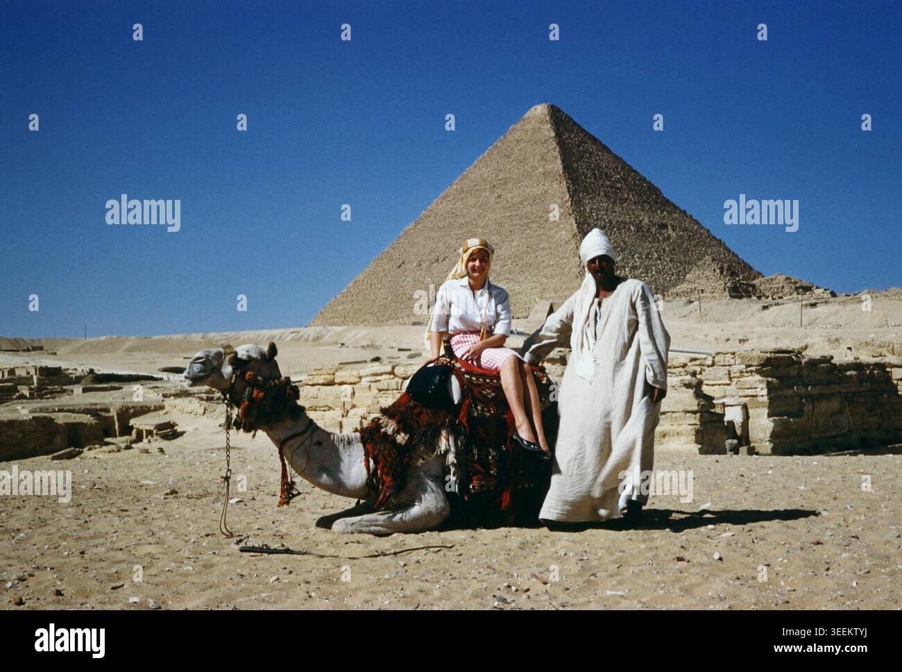 Una turista femminile seduta in alto su un cammello completamente sellato, impegnata in una conversazione con un addetto egiziano locale che indossa una tradizionale veste bianca e fluida galabeya, sullo sfondo di un paesaggio desertico con una grande piramide che si erge dietro di loro. Foto Stock
