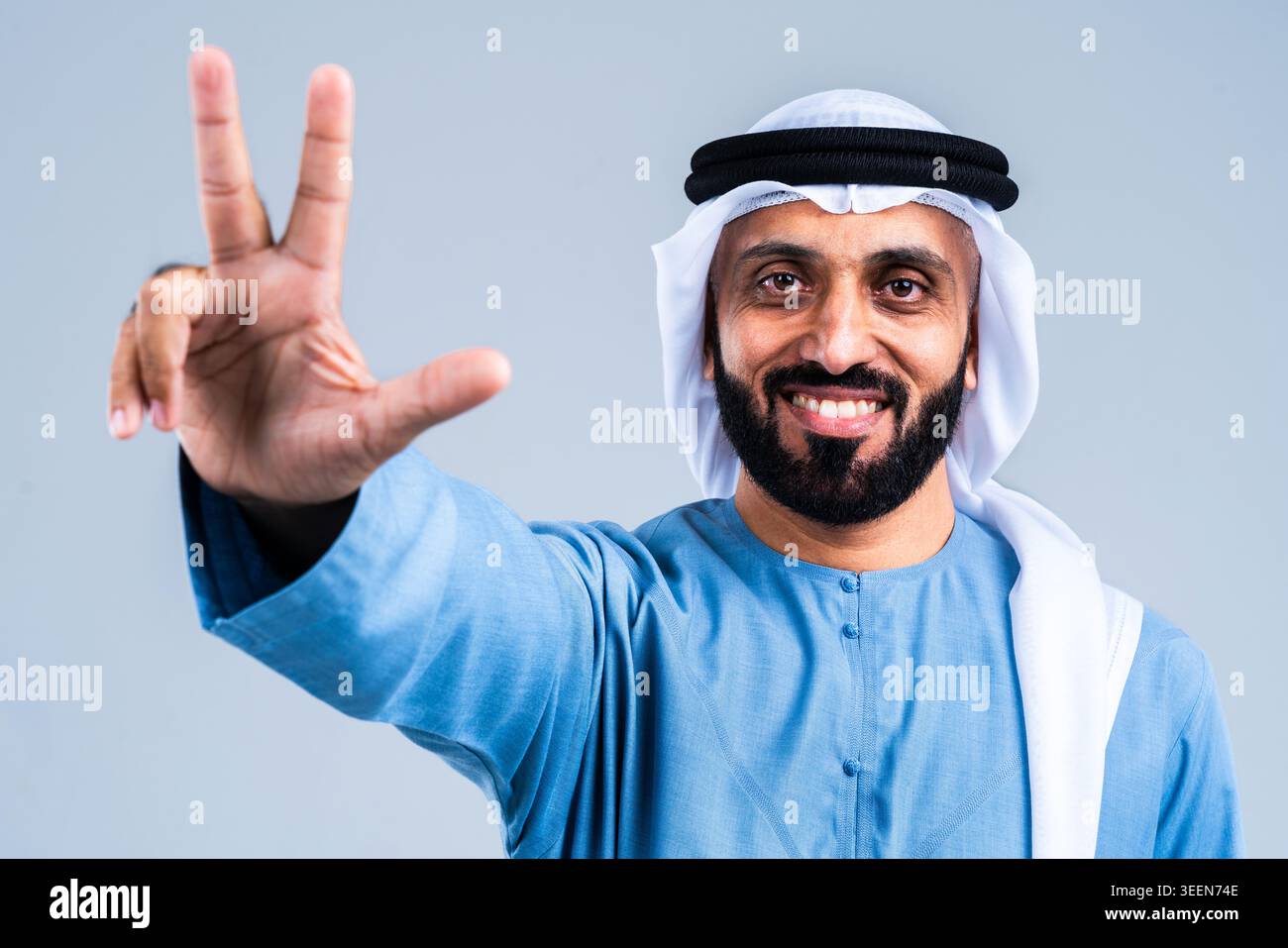 Sorridente uomo barbuto del Medio Oriente che indossa la tradizionale kandura e ghutra degli emirati, alzando la mano, formando l'unico gesto di vittoria d'amore usato Foto Stock
