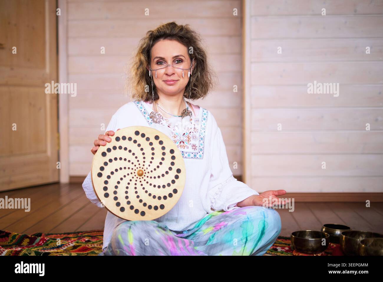Ritratto di un professionista del suono sorridente seduto a gambe incrociate su un tappeto e con un tamburo a cornice in uno studio. Consapevolezza, sollievo dallo stress e. Foto Stock