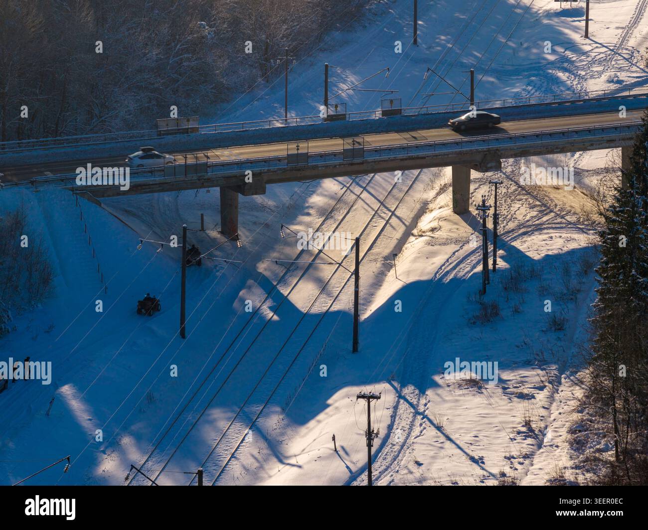Vista aerea di un ponte stradale che attraversa i binari ferroviari innevati con persone e quad in una foresta invernale con lunghe ombre in Lituania. Foto Stock