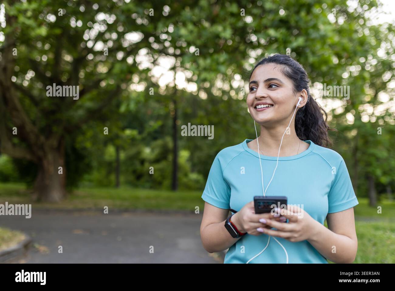 Giovane donna che ascolta la musica tramite auricolari cablati sul suo smartphone, indossa un fitness tracker e una t-shirt sportiva, sorridendo felicemente in un ambiente verde sfocato Foto Stock