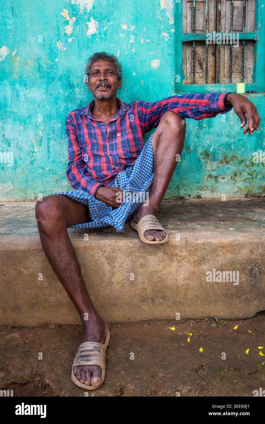 Uomo di mezza età che riposa fuori da una casa tradizionale dai colori vivaci in un villaggio rurale di Orissa, in India Foto Stock