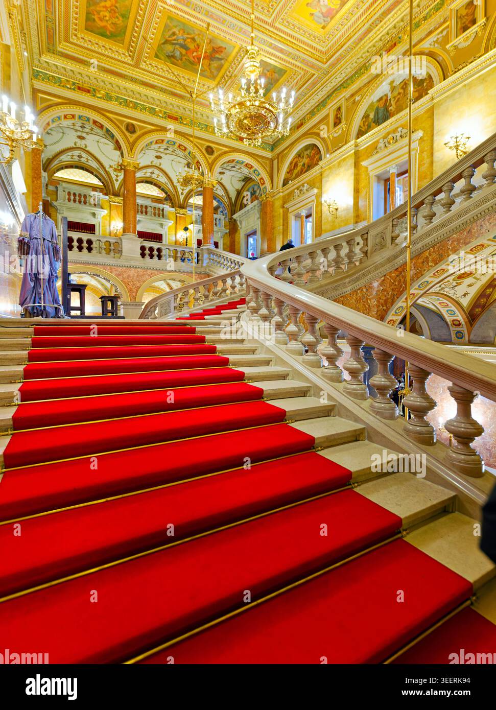 Le persone si spostano sulle scale del tappeto rosso dell'Opera di Stato ungherese. La sala ha luci luminose e disegni decorati sulle pareti. Foto Stock
