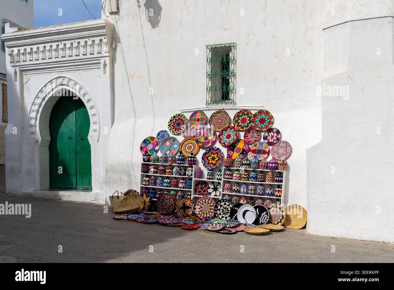 Souvenir colorati esposti su una parete bianca con una porta tradizionale dipinta di verde con una cornice ad arco nella città vecchia di Asilah, in Marocco. Foto Stock