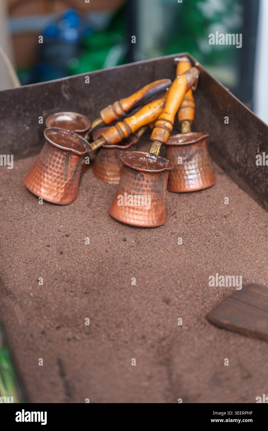 Caffettiere in rame con manici in legno appoggiati su sabbia calda per la preparazione tradizionale della birra. Foto di alta qualità Foto Stock
