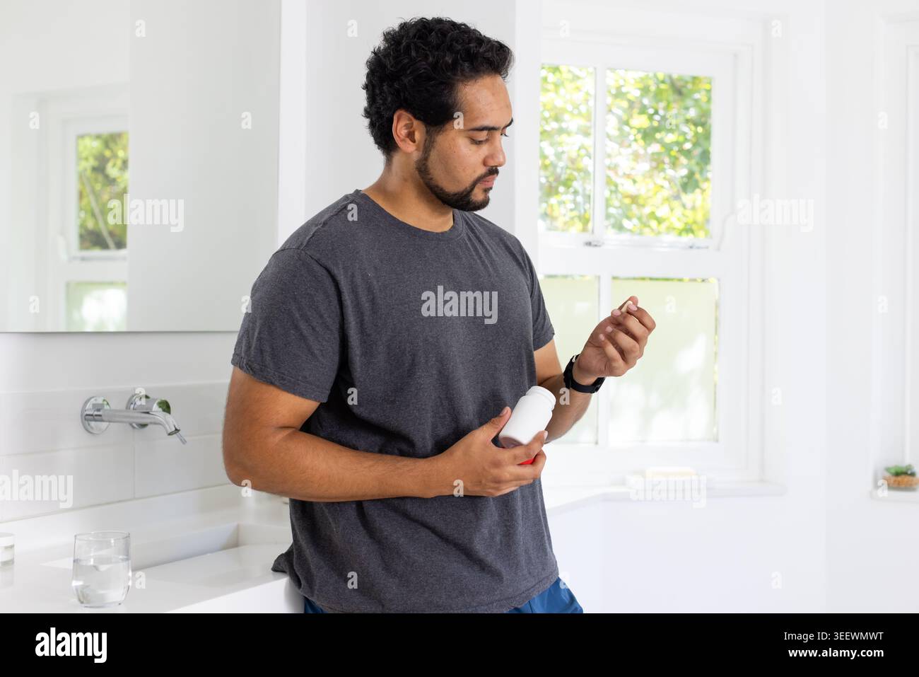 Uomo afroamericano medio adulto in T-shirt grigia e pantaloncini blu, appoggiato al lavandino che regge una bottiglia di pillola Foto Stock