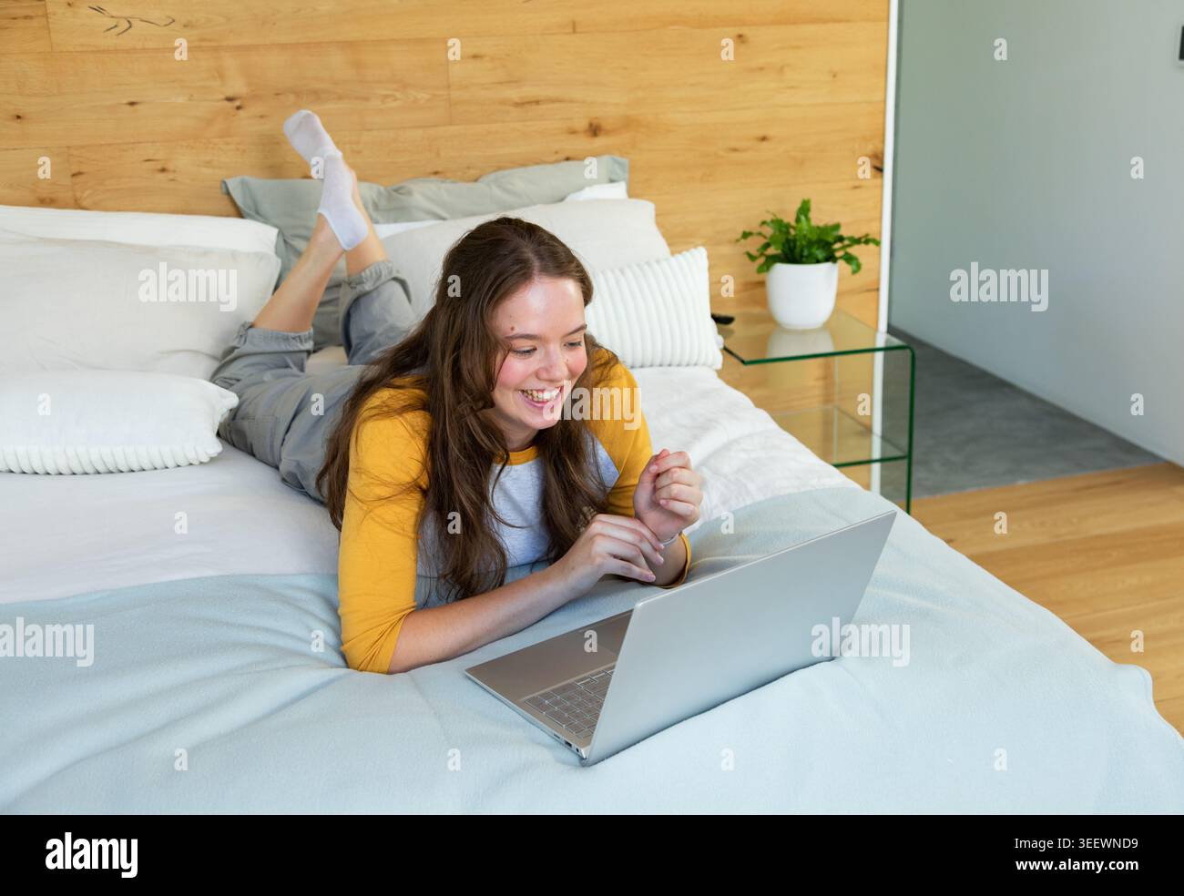 Donna adulta sdraiata sul letto sorridente di un laptop argentato in camera da letto con maniche gialle Foto Stock