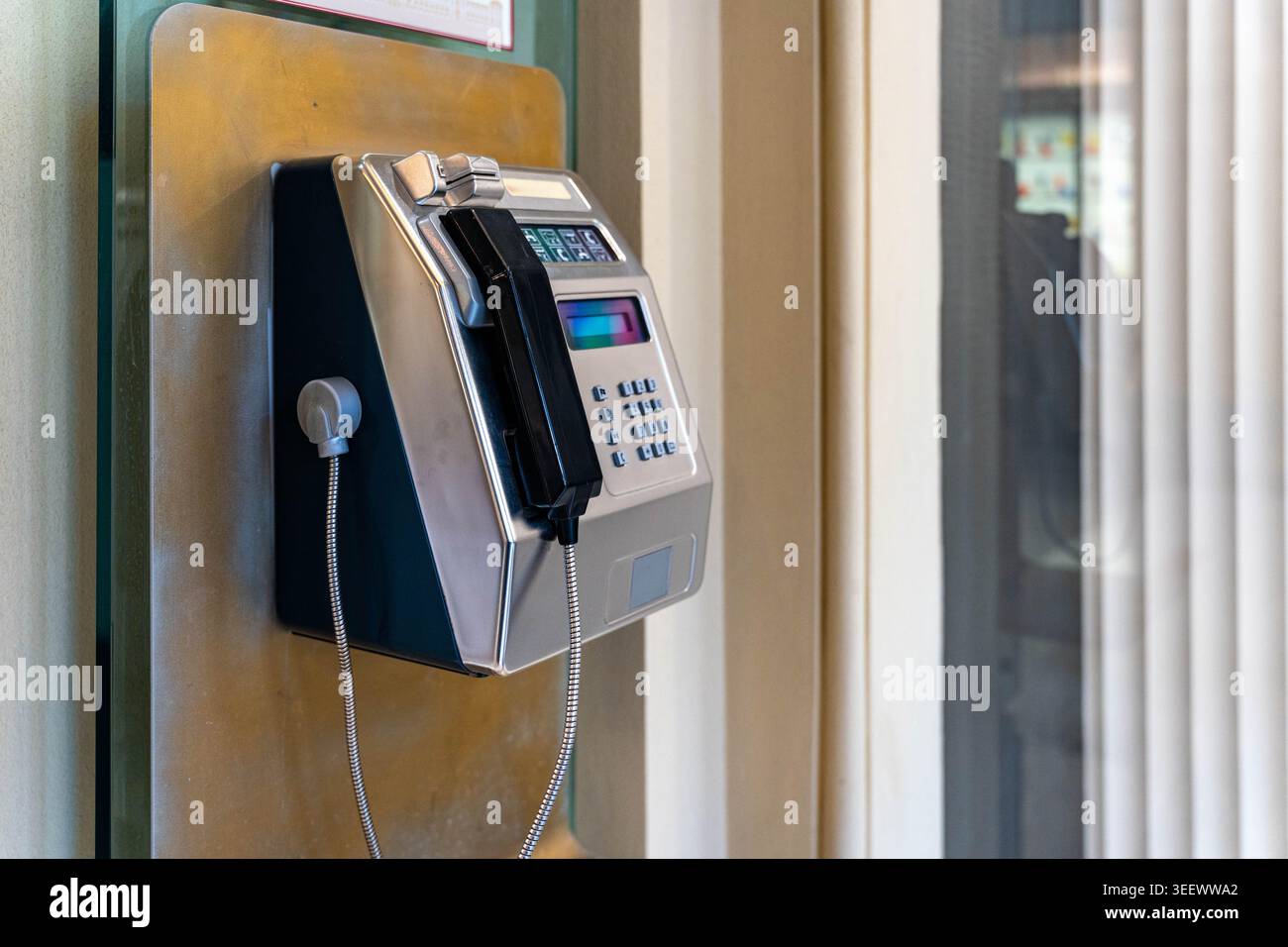 Telefono cellulare vintage montato a parete accanto alla finestra in vetro nella hall tranquilla, rivestimento in metallo lucido e tastiera numerica, cavo ricurvo avvolto verso il basso, slot per monete Foto Stock