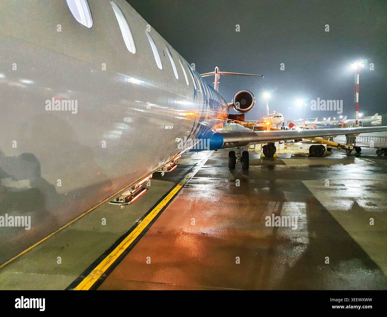 Prima piano ad angolazione bassa della fusoliera di un aereo di linea commerciale di notte su una pista aeroportuale bagnata. L'aereo argentato metallico con accenti blu è Foto Stock