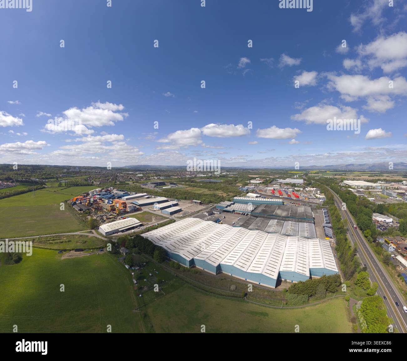 Vista aerea di grandi magazzini industriali ed edifici commerciali circondati da campi verdi sotto un cielo blu con nuvole a Grangemouth, Scozia, Foto Stock