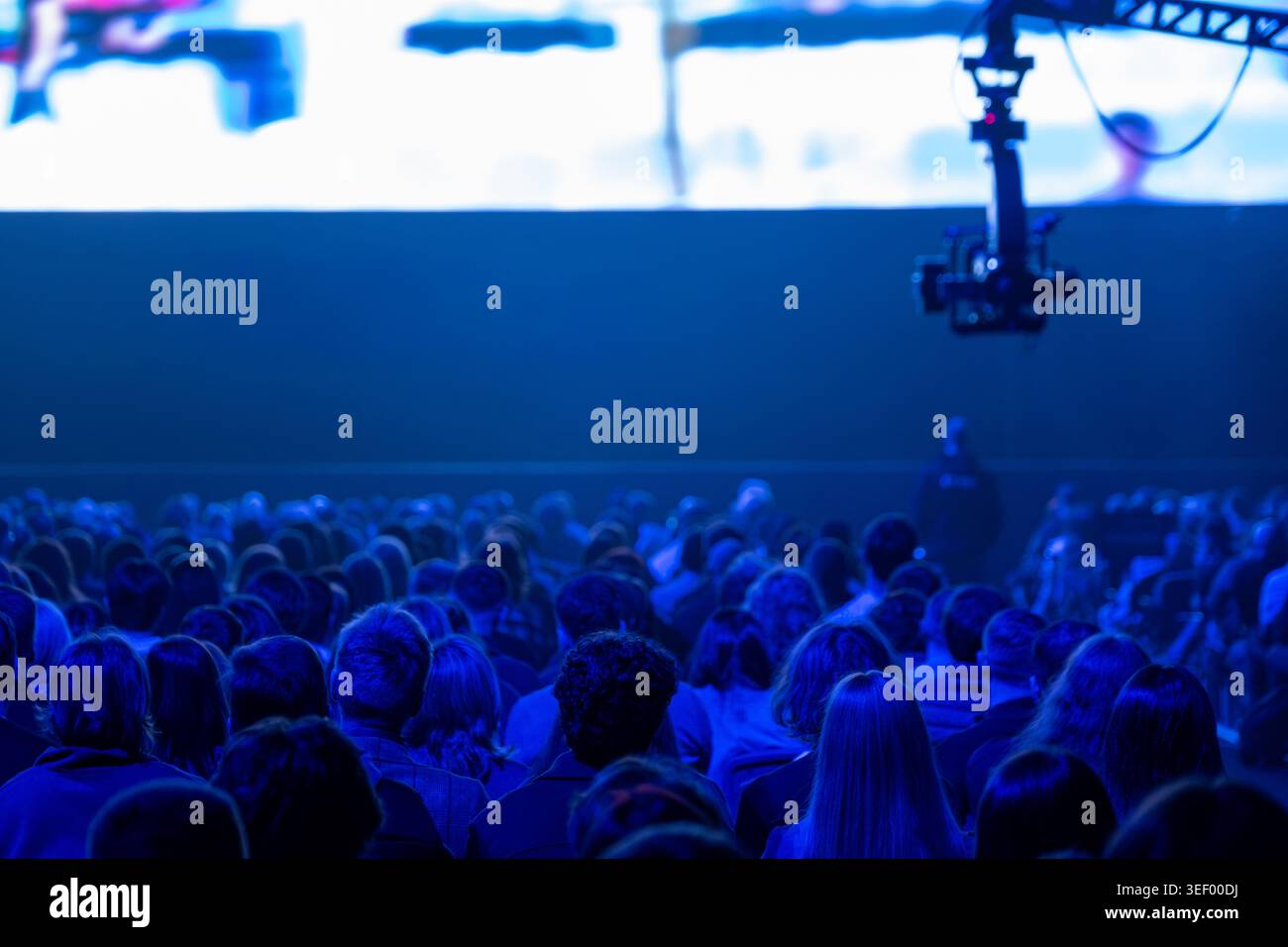 Un pubblico numeroso seduto in una luce blu tenue guardando la presentazione della conferenza sul grande schermo, attrezzatura per telecamere visibile sopra il palco. Foto Stock
