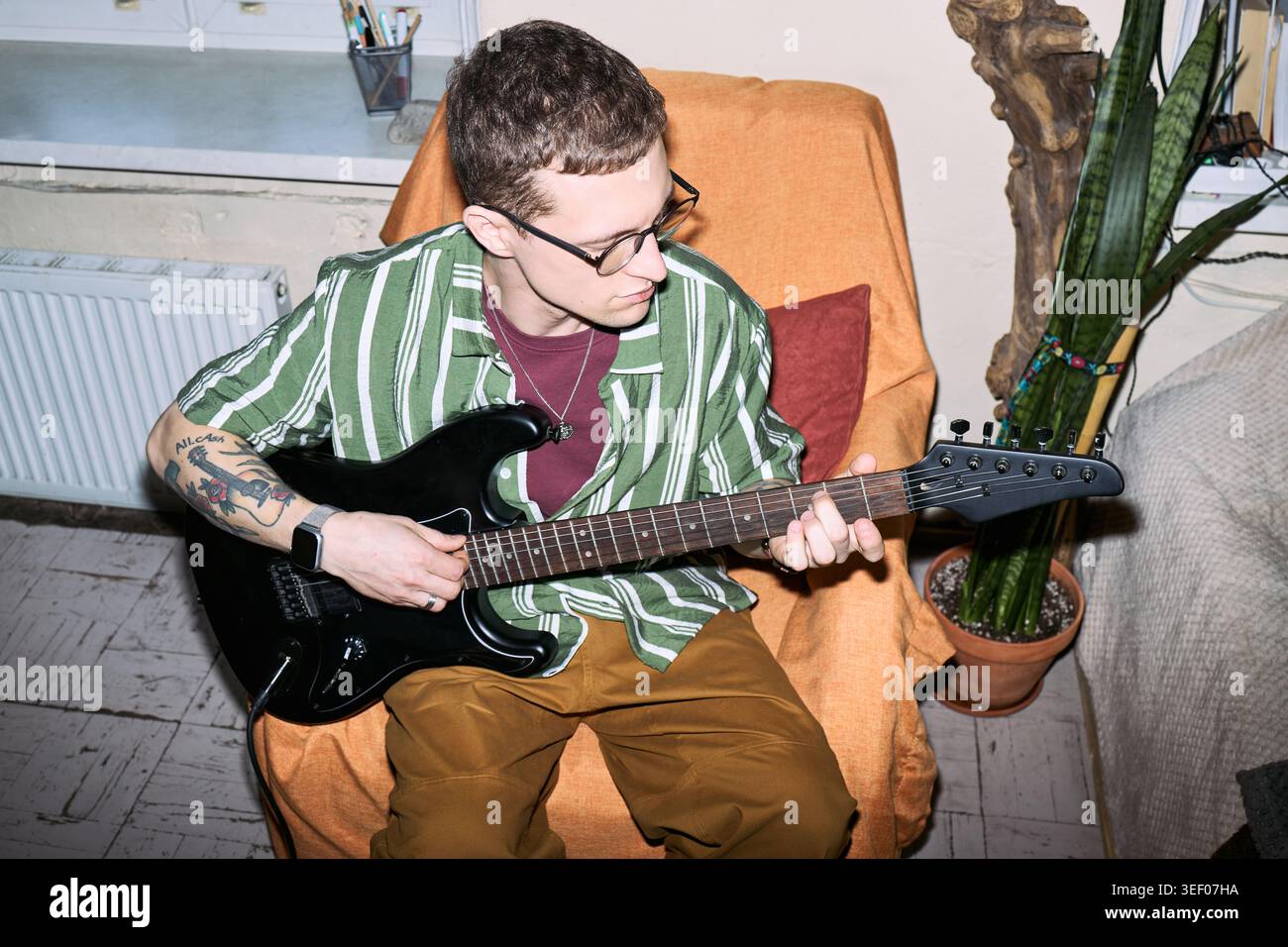 Giovane uomo caucasico adulto che suona la chitarra elettrica mentre è seduto a casa, praticando musica e hobby creativo. Utile per marketing lifestyle, lezioni di musica, concetti per il tempo libero Foto Stock