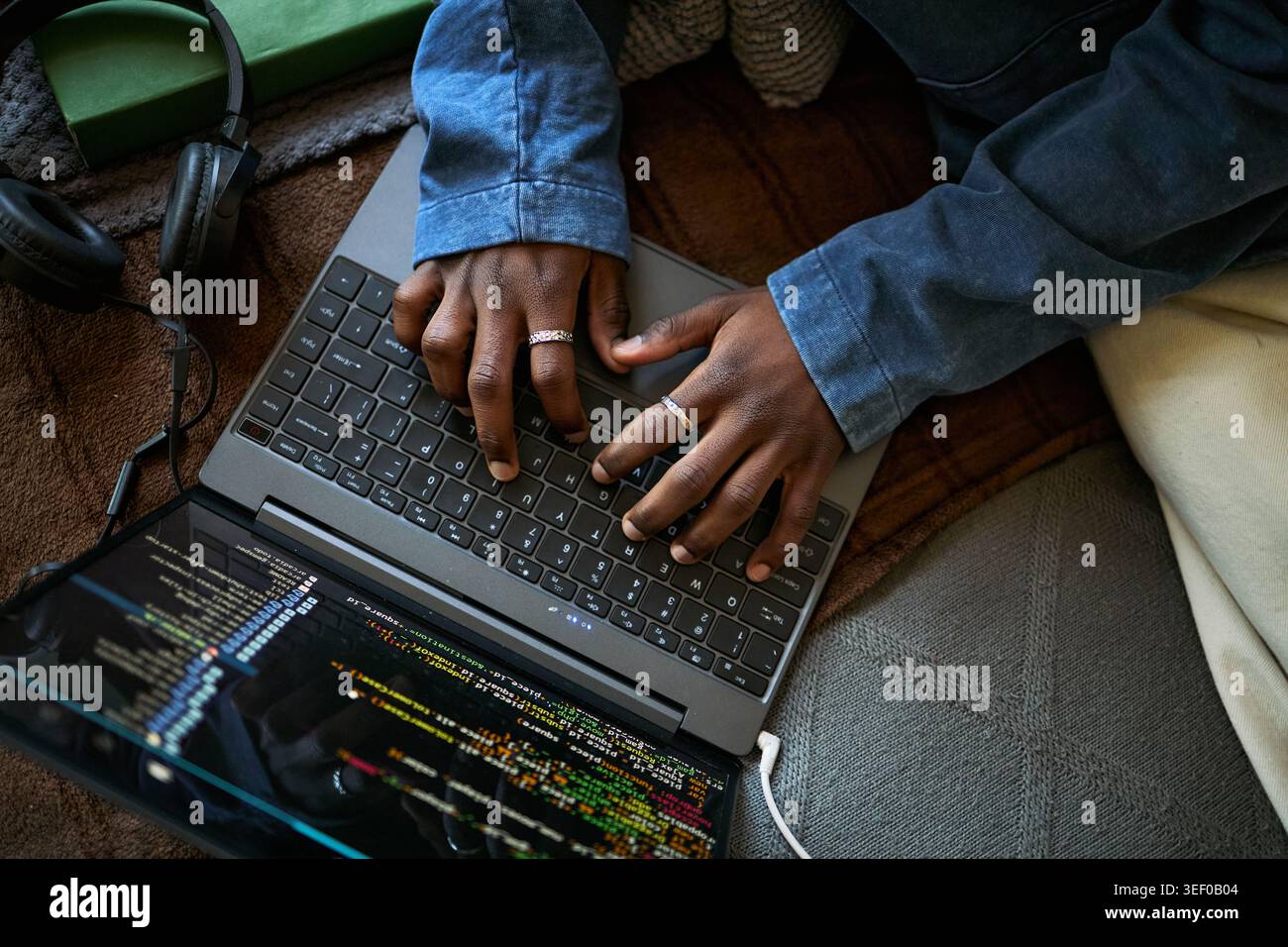 Primo piano di un giovane adulto nero che scrive sul laptop con schermo di codifica e cuffie, lavorando in remoto su progetti software. Ideale per attività di marketing in remoto, programmazione e formazione tecnica Foto Stock