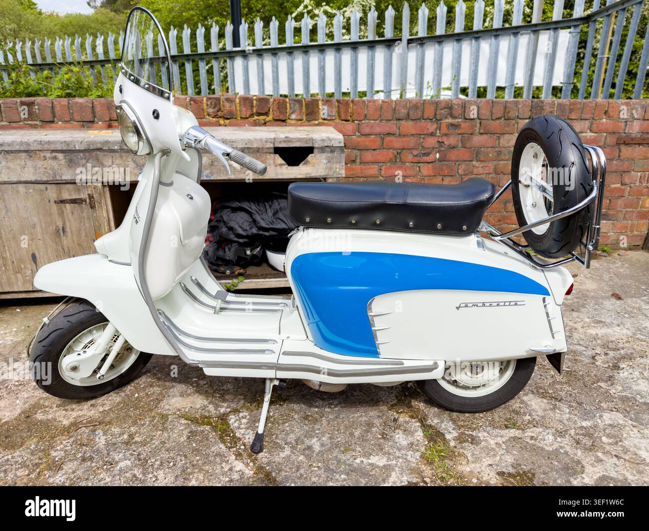 Vista profilo di uno scooter Lambretta TV200 vintage bianco e blu anni '1960 parcheggiato su una superficie in pietra contro un muro di mattoni con parabrezza e ruota di scorta Foto Stock
