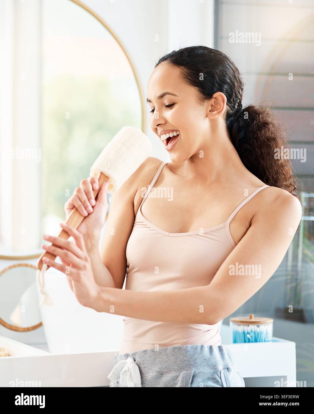 Donna, bagno e canto con loofah al mattino con sorriso, voce e giocosa con benessere allo specchio. Ragazza, spugna e felice con musica, canzone e.. Foto Stock