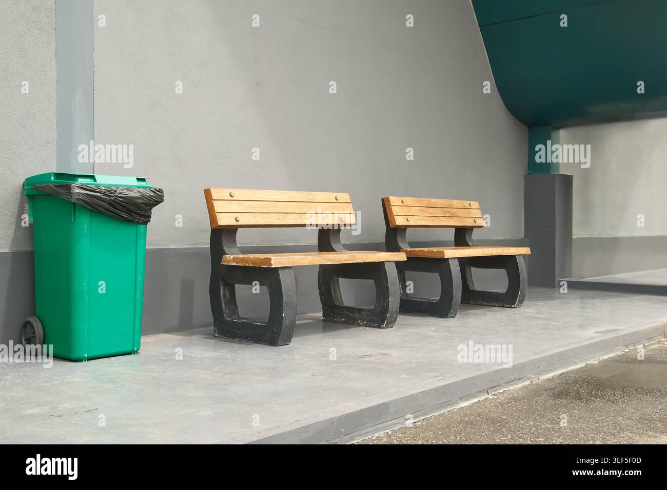panche vuote in legno con supporti in calcestruzzo e cestino in plastica verde, mobili urbani su parete grigia, vista laterale sulla piattaforma Foto Stock