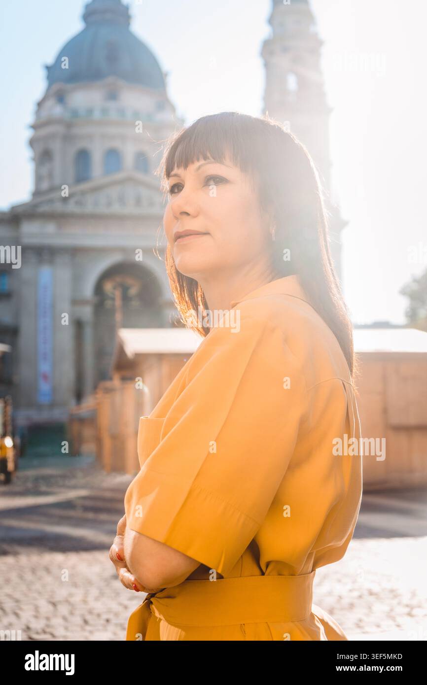 Ritratto di una donna matura e sicura di sé in piedi all'aperto, sotto la calda luce del sole della sera, in una storica città europea. Viaggio da solo, indipendenza e concetto di stile di vita urbano moderno. Foto Stock