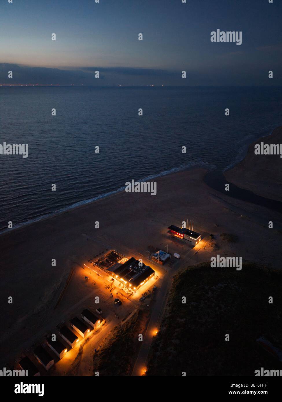 Vista aerea notturna dei Beach club e dei caffè illuminati su una costa sabbiosa del Mare del Nord. Luci calde si illuminano contro l'oceano scuro e il cielo crepuscolo in questo sc Foto Stock