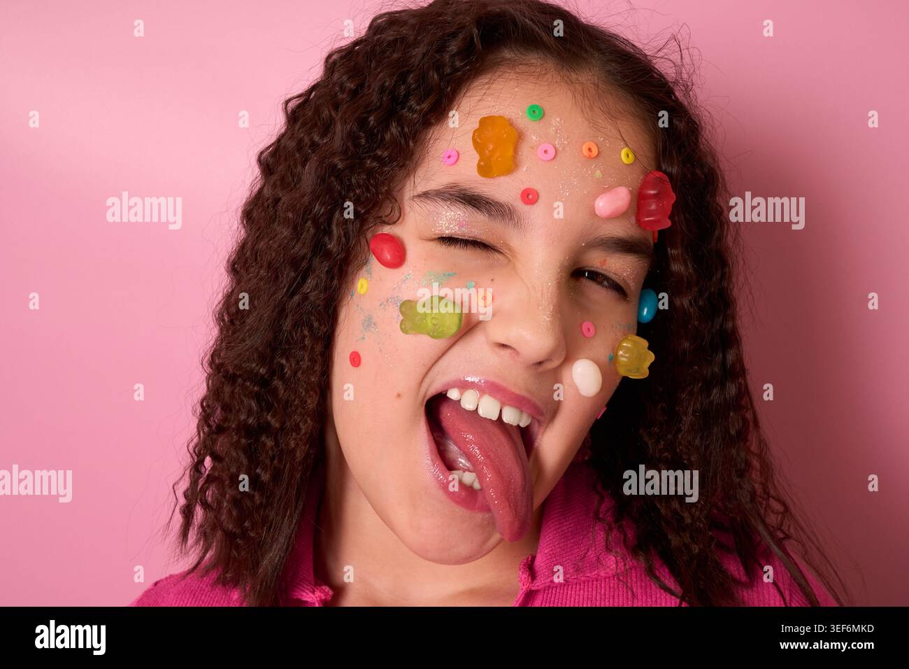 Playful Girl con coloratissimi adesivi di caramelle sul viso che creano espressioni divertenti su sfondo rosa Foto Stock