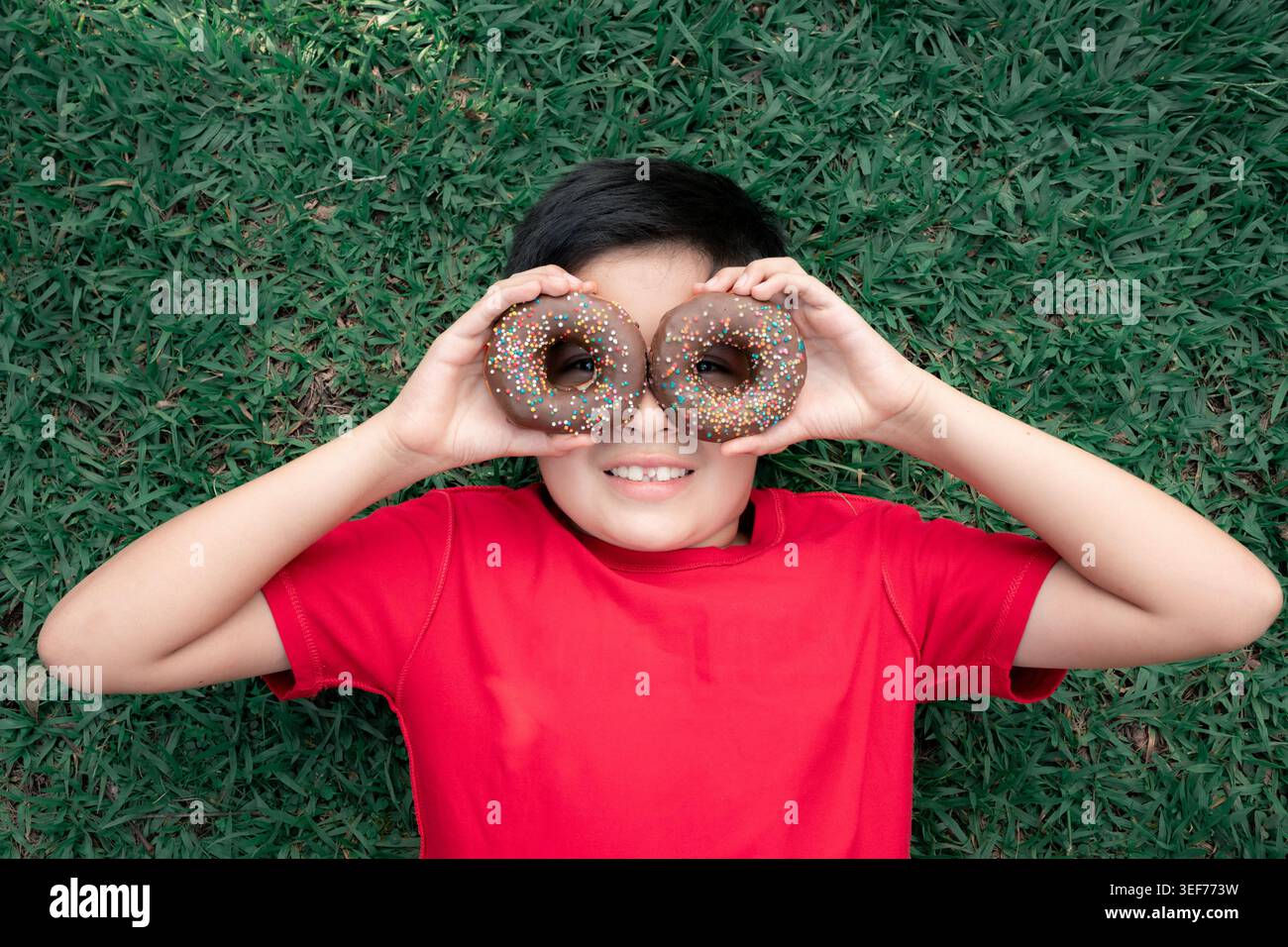 Ragazzo disteso su erba verde fresca, con due ciambelle al cioccolato con spruzzi colorati sugli occhi, sorridendo e giocando all'aperto in una giornata di sole in un r Foto Stock