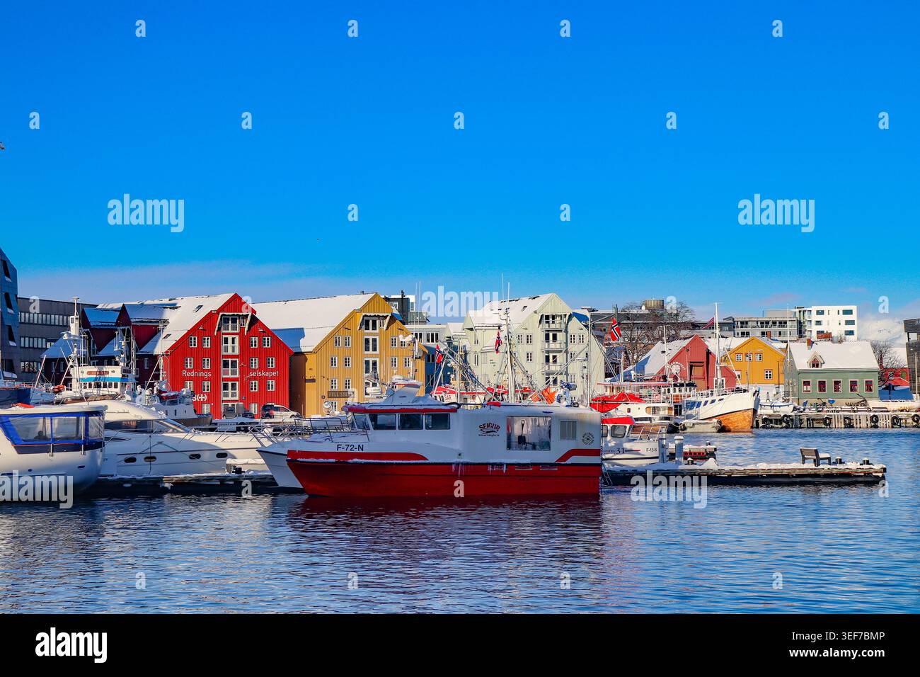 Fila di edifici storici in legno di colore rosso e giallo di fronte al porto di Tromso, con barche ancorate e neve sotto un cielo blu profondo, la Norvegia. Foto Stock