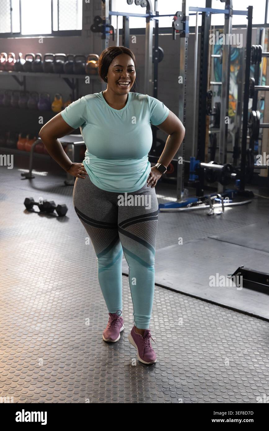 Donna afro-americana sorridente che indossa leggings grigi color acqua in palestra vicino al portamonete Foto Stock