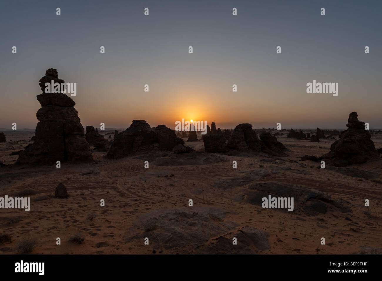Sagome di cairn di pietra e pinnacolo di arenaria che circondano il sole splendente all'orizzonte al tramonto del deserto, regione di Alula, Arabia Saudita Foto Stock