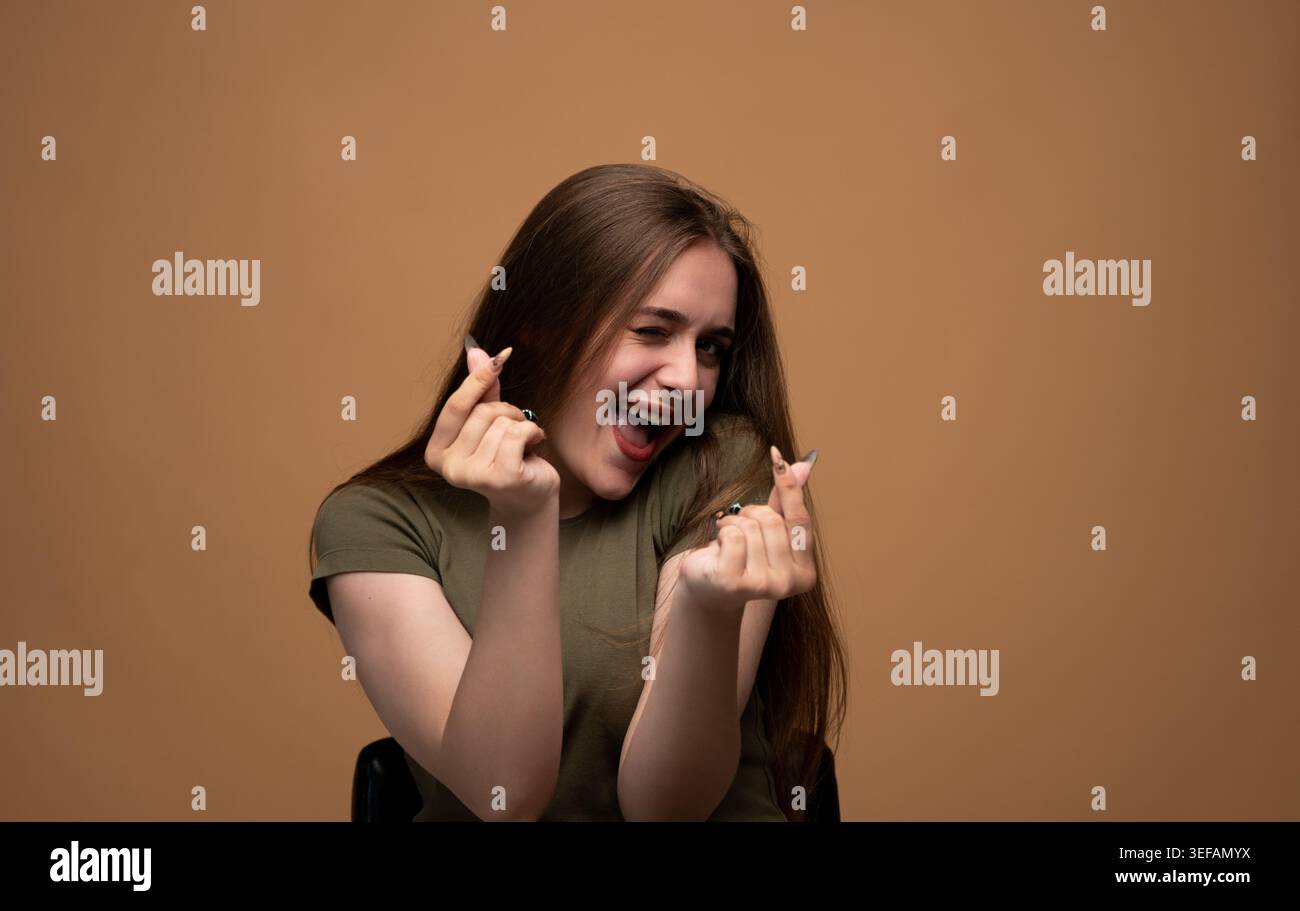 Allegra giovane donna che si alza, ride e fa gesti coreani con le dita sullo sfondo marrone dello studio Foto Stock