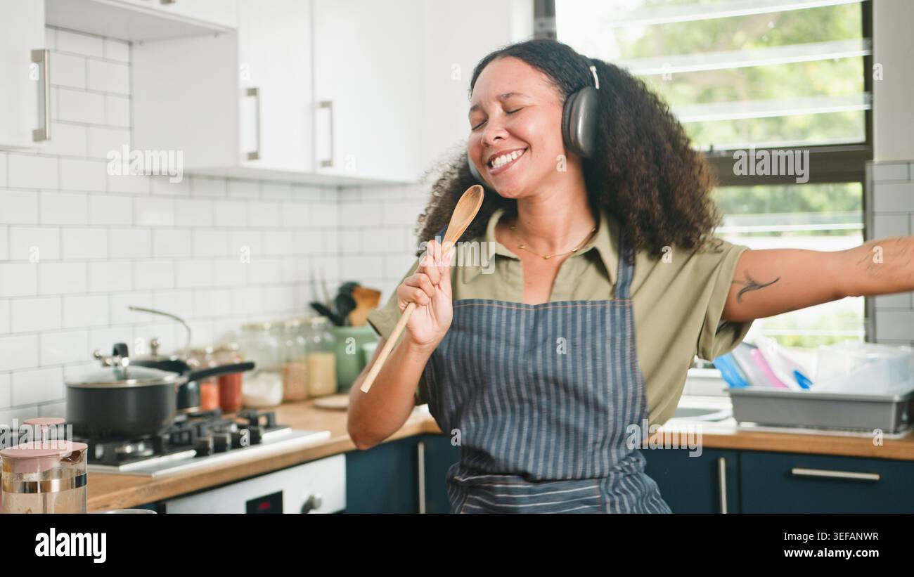 Donna allegra che canta e balla in cucina mentre cucina con le cuffie e il cucchiaio di legno Foto Stock