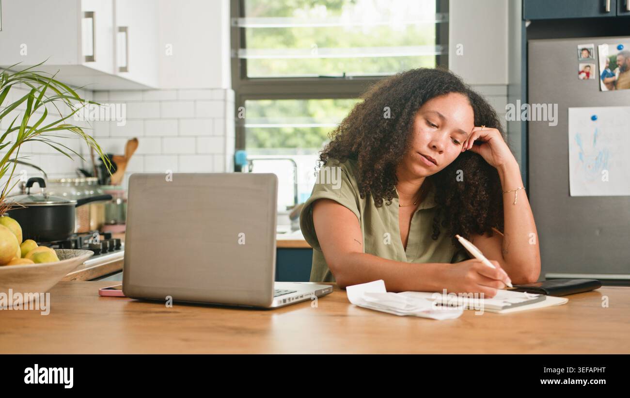 Donna stressata al tavolo da cucina con notebook che lavora su bollette e finanze mentre scrive appunti Foto Stock