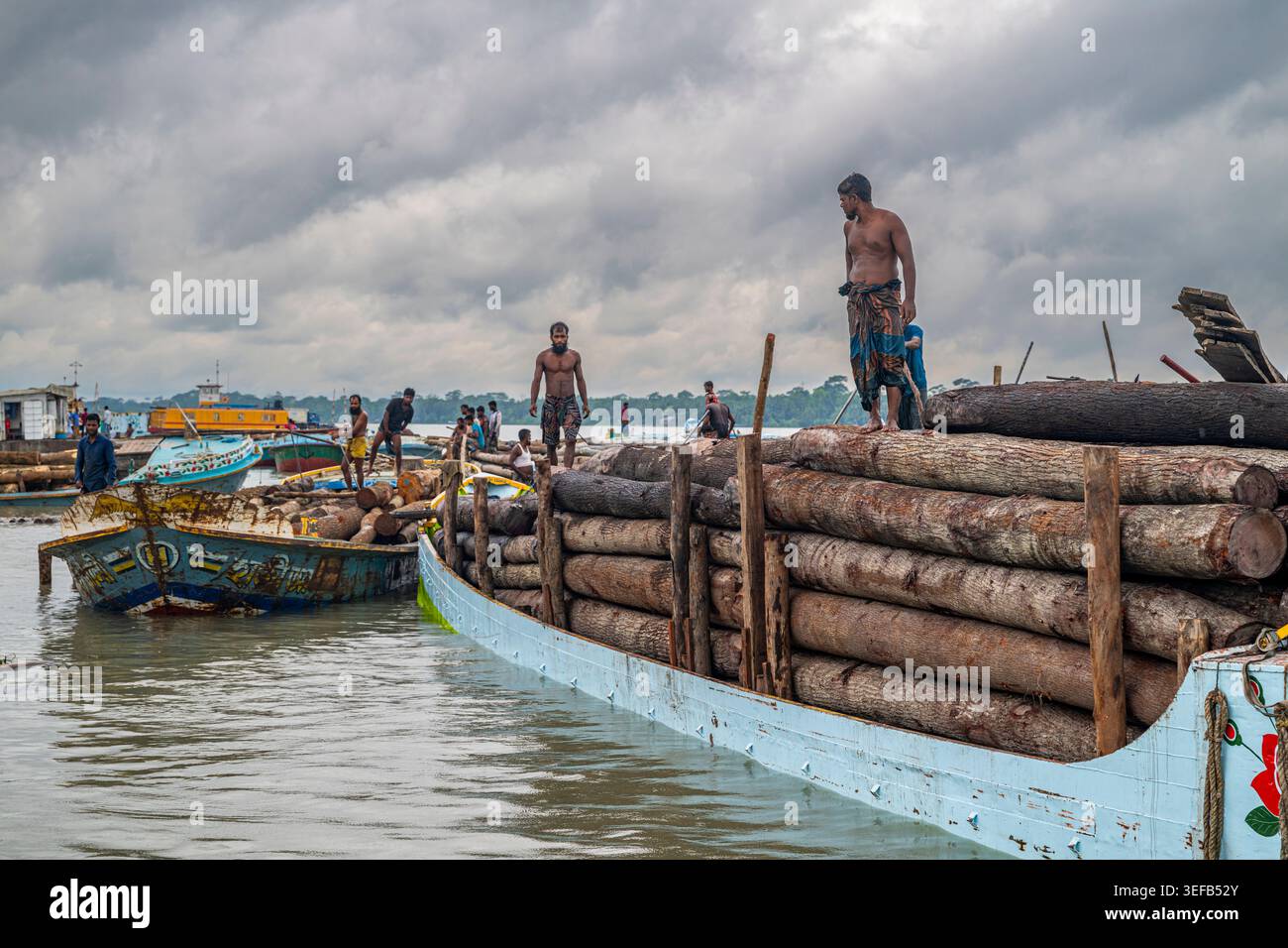 I lavoratori trasportano tronchi in un mercato del legno lungo i canali d'acqua a Barishal, in Bangladesh, durante una giornata nuvolosa Foto Stock