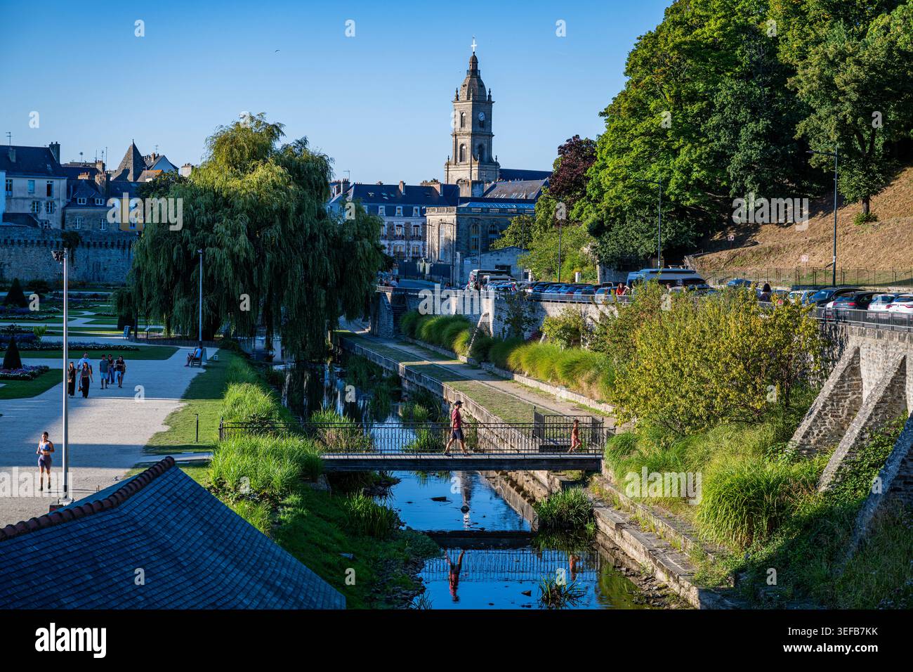 I visitatori camminano vicino al fiume e agli edifici in una giornata di sole in una città francese con una storica torre dell'orologio e alberi verdi Foto Stock