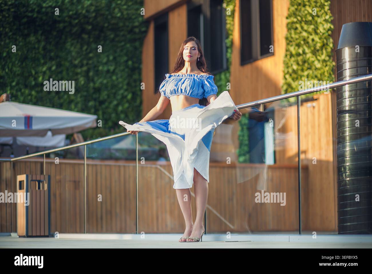 Giovane donna sicura di sé con i capelli lunghi si posa in elegante gonna e top blu e bianco. Si erge graziosamente su un balcone moderno con luce calda del sole. Backgrou Foto Stock