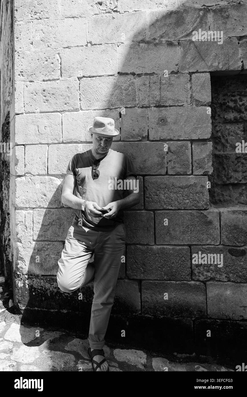 Fotografia di strada monocromatica con un uomo alla moda che si appoggia alla vecchia architettura nel vicolo mediterraneo della città. Foto Stock