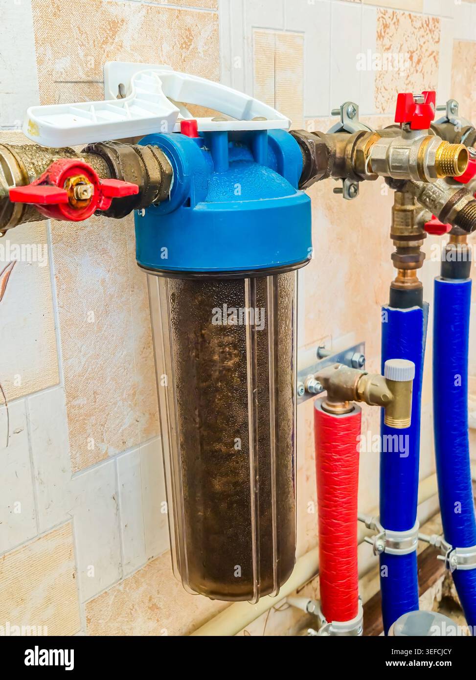 Un alloggiamento del filtro dell'acqua trasparente ostruito riempito di sedimenti e detriti, collegato a tubazioni e valvole in un ambiente residenziale Foto Stock