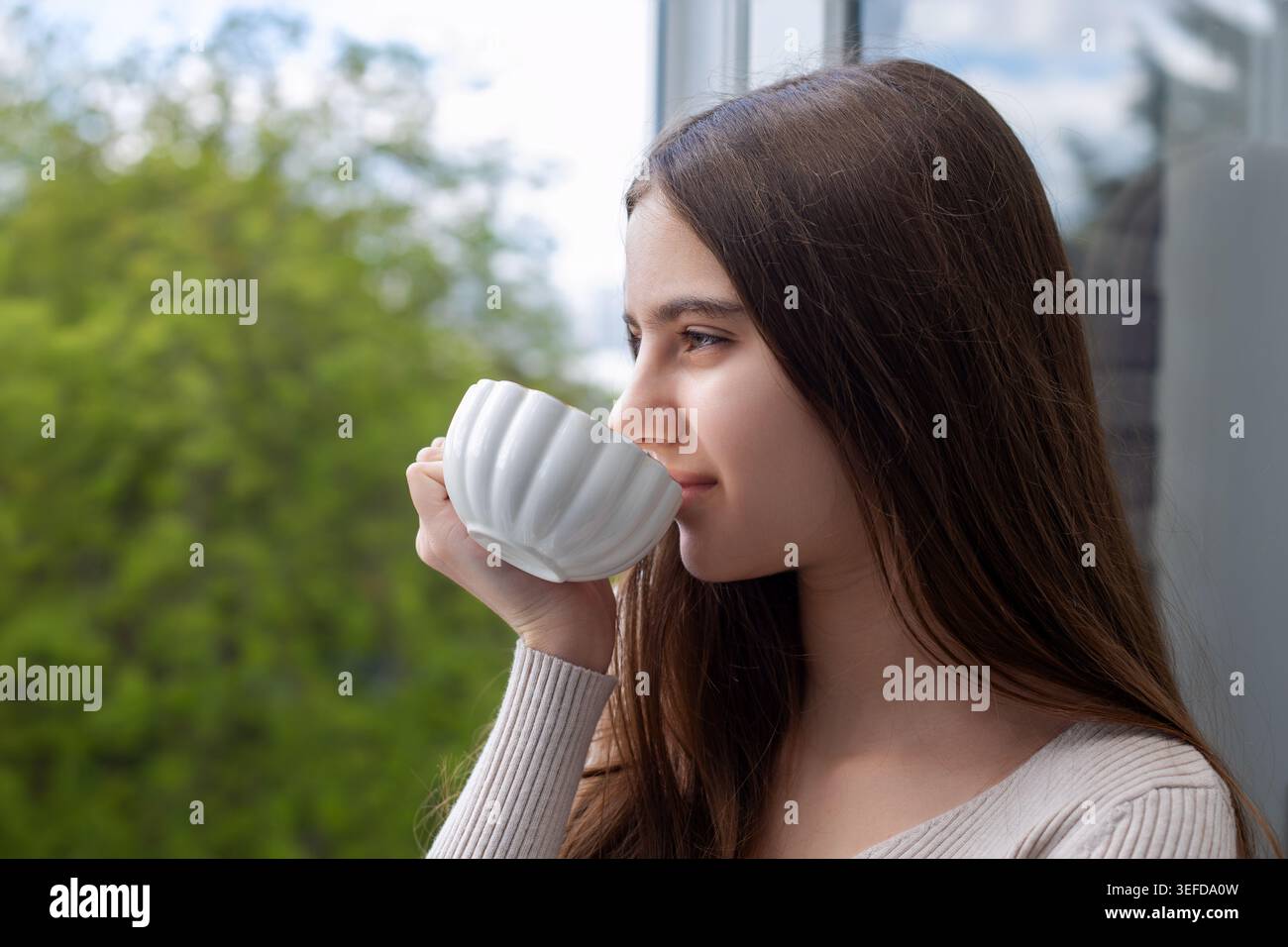 Giovane donna che beve il tè vicino a Window. Giovane donna che beve da una tazza bianca accanto a una finestra aperta, godendosi un momento di calma con la natura verde all'aperto. Foto Stock