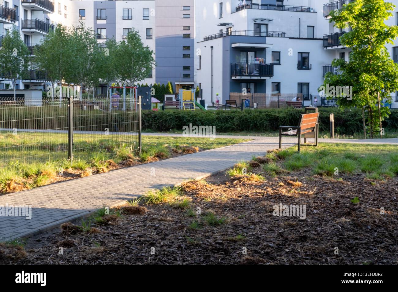 Il sentiero pedonale pavimentato passa accanto a una panchina del parco e a uno spazio verde recintato all'esterno dei moderni edifici residenziali. Foto Stock