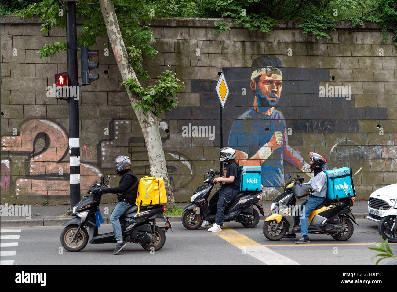 I motociclisti che viaggiano in scooter passano davanti a un grande murale di un uomo su un muro di pietra. Foto Stock