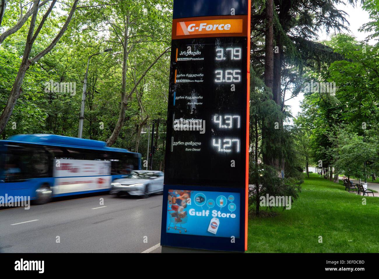 Un alto cartello del prezzo del carburante mostra vari prezzi del carburante in un parco verde con alberi e un autobus che passa. Foto Stock