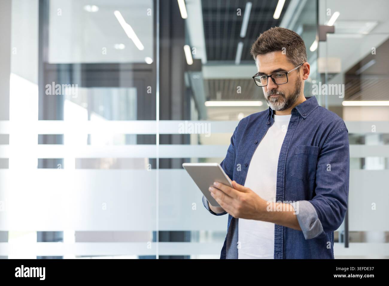 Uomo d'affari con il barbuto che indossa occhiali e una camicia in denim che si impegna attivamente con un tablet digitale, gestisce gli affari e si connette in un ambiente d'ufficio contemporaneo Foto Stock