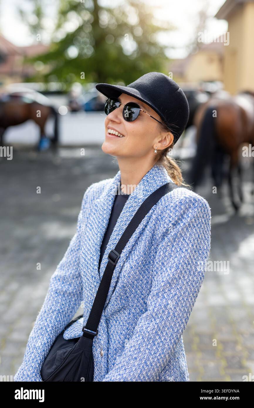 Elegante donna sorridente che indossa occhiali da sole e berretto vicino ai cavalli nella stalla di campagna. Donna elegante che gode di una giornata di sole all'aperto con relax Foto Stock
