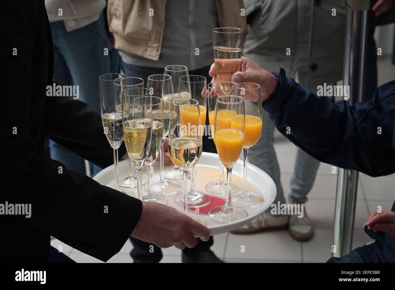 Cameriera che serve un vassoio di champagne e succo d'arancia ad un ricevimento di benvenuto, persone in abiti informali che vi stanno intorno e portano i bicchieri, selezionate Foto Stock
