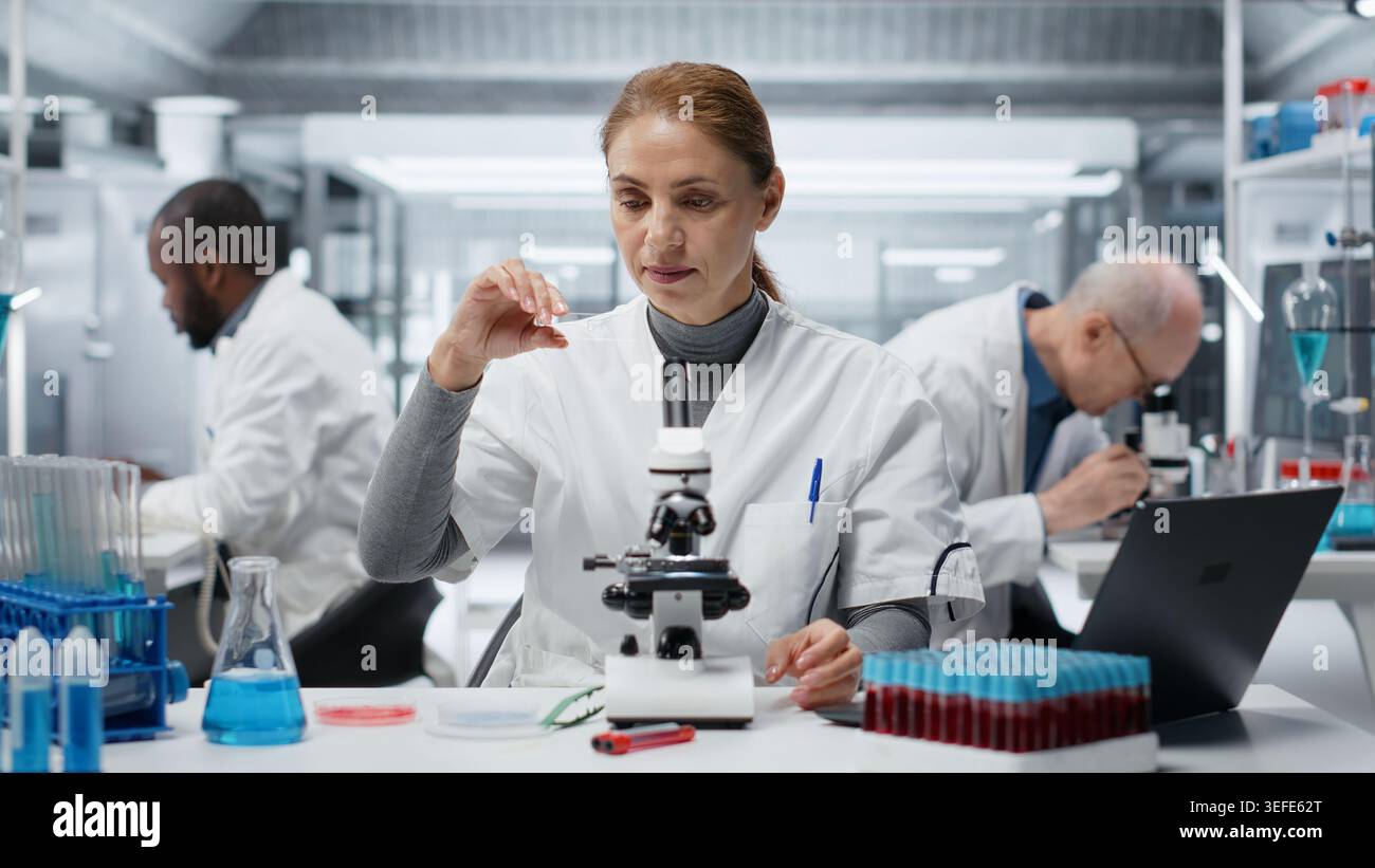 Tecnico istologico in laboratorio che ispeziona vetrini sottili per verificare la presenza di difetti strutturali o impurità. Donna in laboratorio che esegue il controllo visivo della qualità utilizzando un microscopio composto, telecamera B. Foto Stock