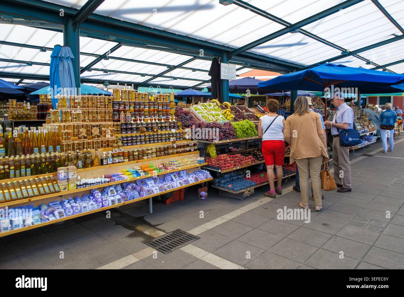 Turisti che navigano in un mercato all'aperto pieno di miele, olio d'oliva, lavanda e frutta fresca sotto una struttura con tetto in metallo in Croazia, Europa Foto Stock