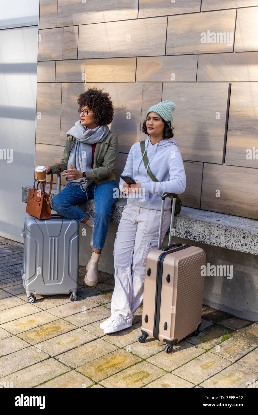 Due amiche in abiti da viaggio aspettano fuori da un edificio moderno con una panchina bassa e valigie Foto Stock