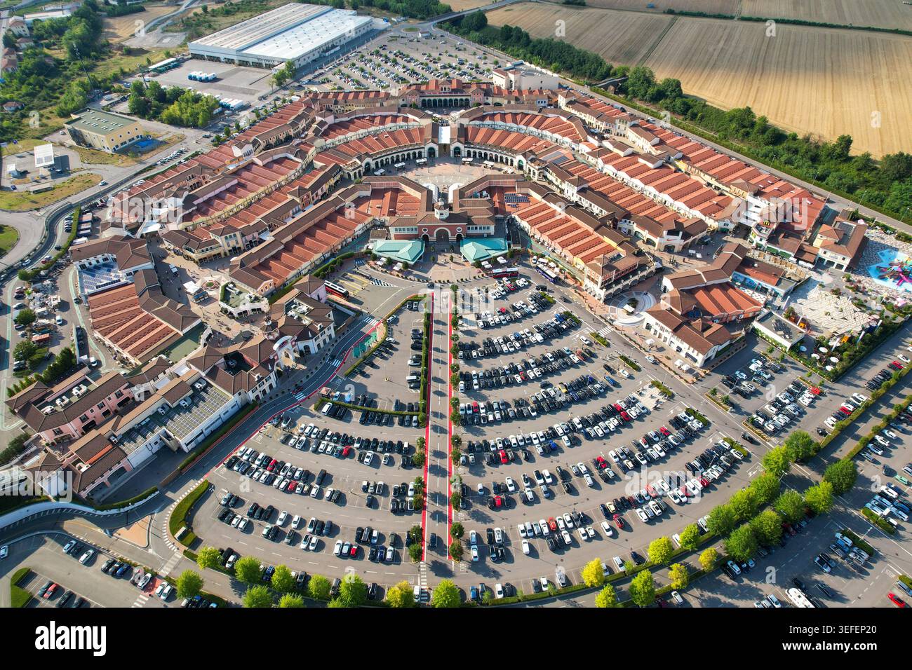 Vista aerea del Designer Outlet Serravalle, caratterizzato dalla sua caratteristica architettura circolare e dai tetti di tegole rosse, circondato da grandi aree parcheggio a Ser Foto Stock